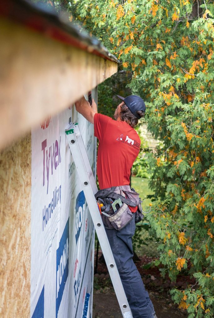 Adult male construction worker wearing a red shirt repairs roof siding outdoors with green trees in background.