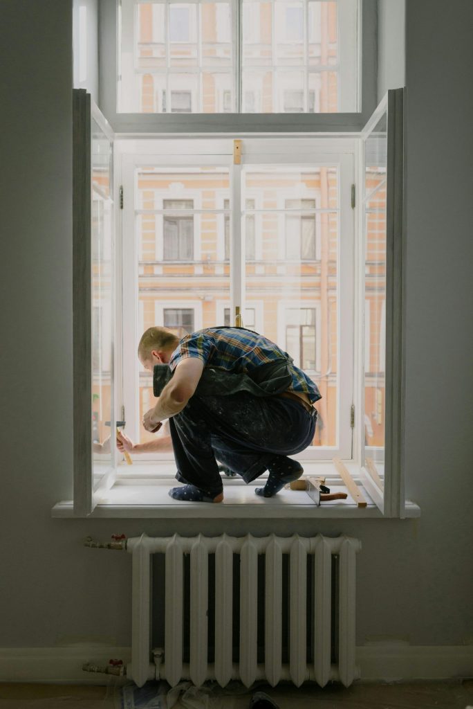 Full body of unrecognizable handyman on windowsill with instruments assembling window while working in room with battery during renovation works